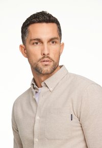 Camicia beige con colletto, taschino sul petto e tessuto strutturato. Cuciture sottili e un piccolo marchio sul taschino.