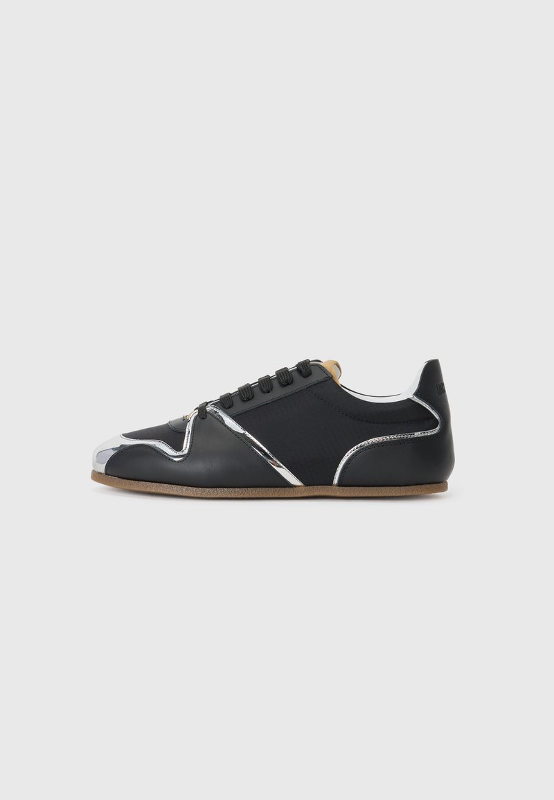 Schwarzer Sneaker mit niedrigem Schaft, silbernen Akzenten, schwarzen Schnürsenkeln, braunem Innenfutter und dünner brauner Sohle vor schlichtem hellgrauem Hintergrund.