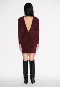 Mini-jurk in bordeaux met glans, lange mouwen en een diepe V-rug. Gecombineerd met zwarte, kniehoge, puntige laarzen. Soepele textuur en nauwsluitend ontwerp.