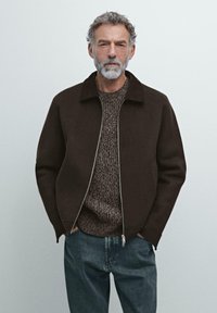 Giubbotto marrone con zip e colletto alto sopra un maglione marrone a trama e jeans blu. La barba dell'uomo si abbina al suo complesso palette neutra.