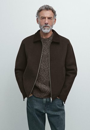 Veste brune zippée avec col montant sur un pull brun texturé et un jean bleu. La barbe de l'homme complète sa palette neutre globale.