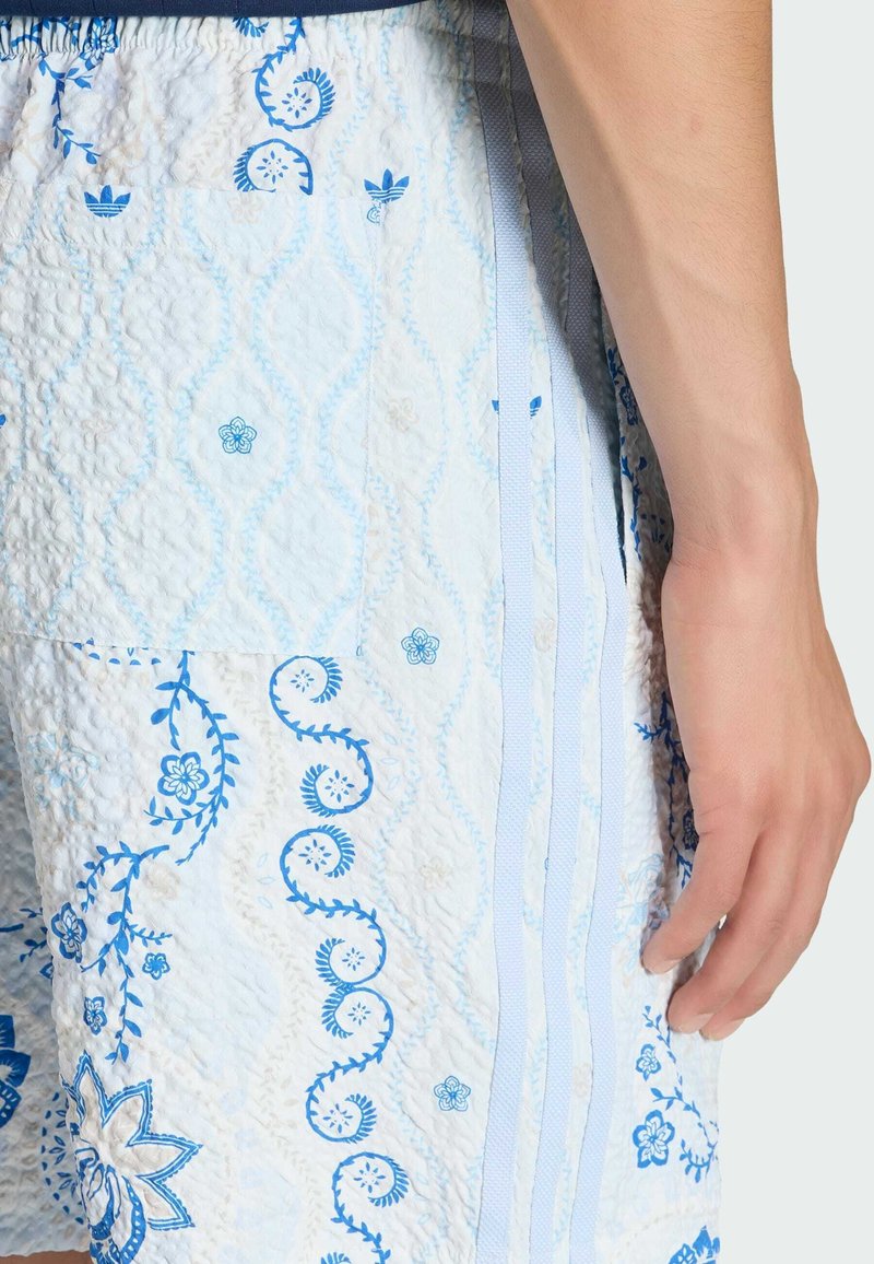 Rechterhand rust naast lichtblauwe shorts met een doorgestikte textuur en blauwe bloemen- en krulpatronen.