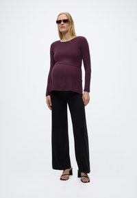 Έγκυος γυναίκα που φοράει μπλούζα μακριά μανίκια σκούρου μωβ, μαύρο παντελόνι με φαρδιά πόδια, μαύρες πλατφόρμες με τακούνι και σκούρα ορθογώνια γυαλιά ηλίου σε λευκό φόντο.