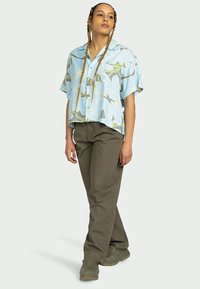 Cămașă bleu deschis cu mânecă scurtă, cu un model floral, asortată cu pantaloni verzi măslinii și încălțăminte gri. Modelul stă în profil pe un fundal neutru.
