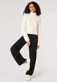 Maglione a collo alto a coste color crema e pantaloni cargo neri a gamba larga con cerniere, abbinati a sneaker bianche, indossati da un modello in una posa casual.