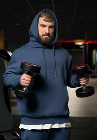 Felpa blu con cappuccio, che tiene due manubri neri con dettagli rossi. Tessuto liscio e taglio atletico, indossata sopra una camicia bianca.