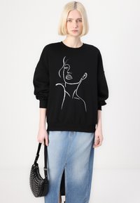 Hanorac negru cu ilustrație albă a unui chip, cu mâneci lungi și manșete elasticizate, asortat cu o fustă din denim cu slit înalt și o poșetă neagră cu studs.