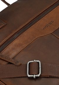 Brown leather backpack with a zippered compartment, fabric strap, metal buckle, and embossed logo detail near the zipper. Textured surface.