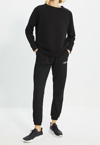 Schwarzer Sportpullover mit Raglanärmeln kombiniert mit schwarzen, schmal geschnittenen Jogginghosen mit Seitenstreifen und besticktem Text. Kombiniert mit schwarzen Turnschuhen.