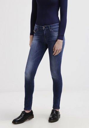 Slank pasform blå jeans båret med sorte læder snørestøvler og en langærmet navy top, stående mod en ensfarvet lys baggrund.
