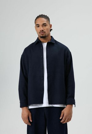 Marineblauw overhemd met knopen in een losse pasvorm, voorzien van een kraag, lange mouwen en een effen textuur. Gestyled over een wit T-shirt, gedragen met donkere jeans.