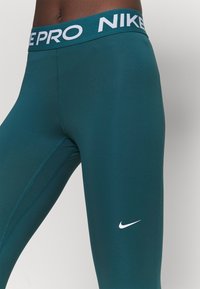 Tyrkysové legíny Nike Pro s bílým logem na pase a po straně, vyrobené z hladkého elastického materiálu, s plochými švy pro pohodlí.
