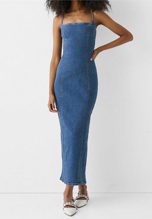 Woman wearing a fitted, sleeveless blue denim maxi dress with thin straps and white pointed-toe flats with black accents.