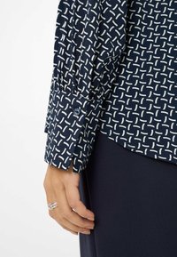 Hand met zilveren ringen naast de manchet van een navyblauw gestreept overhemd met lange mouwen en donkere marineblauwe broek tegen een witte achtergrond.