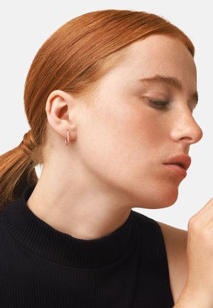Boucle d'oreille créole en or rose portée par un modèle aux cheveux roux. La boucle d'oreille présente un design épuré et minimaliste ainsi qu'une texture lisse et subtile.