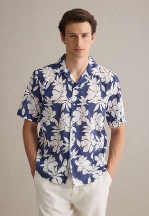 Jonge man met een blauw T-shirt met korte mouwen met grote witte bloemmotieven en witte broek, staand met handen in de zakken.