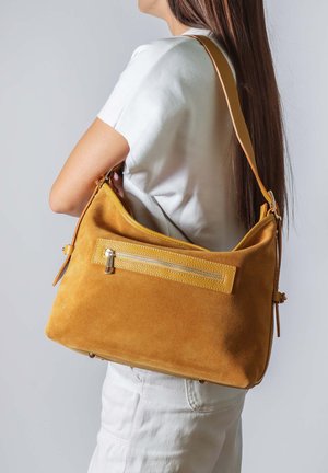 Donna con abiti bianchi che porta una grande borsa a spalla in suede marrone chiaro con tasca frontale con cerniera, su sfondo neutro.