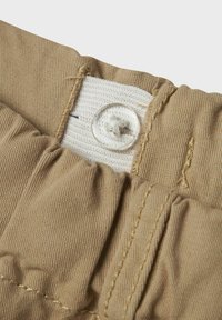 Tissu beige avec une surface texturée, présentant une bande élastique blanche et un bouton en plastique transparent pour le maintien. Gros plan sur les détails de couture.