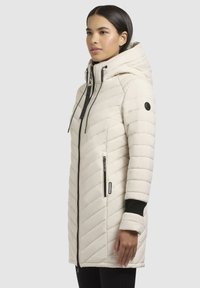 Kvinde iført en mid-length kremfarvet, kviltet vinterjakke med hætte, sort lynlås og sorte håndledsmanchetter, stående mod en ensfarvet baggrund.