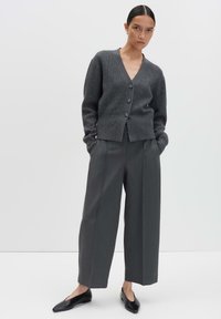 Donna in piedi con le mani in tasca, che indossa un cardigan grigio scuro con bottoni, pantaloni grigi larghi e scarpe nere a punta.