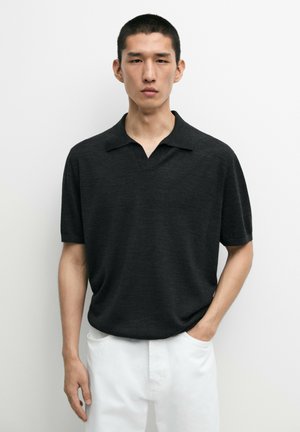 Jeune homme portant un polo noir à manches courtes et un pantalon blanc, debout devant un fond clair uni.