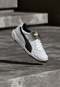 Valkoinen Puma King -jalkapallokenkä mustalla raidalla ja ruskealla pohjalla leijuu betoniporrasaskelmien yläpuolella ulkoilmassa.