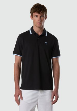 Uomo che indossa una polo nera con rifiniture azzurro chiaro e bianche sul colletto e sulle maniche, abbinata a pantaloni bianchi su uno sfondo semplice.