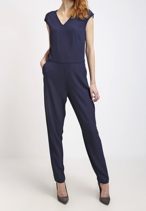 Frau trägt einen marineblauen ärmellosen Overall mit V-Ausschnitt und Seitentaschen, kombiniert mit grauen High Heels, steht vor einem schlichten weißen Hintergrund.