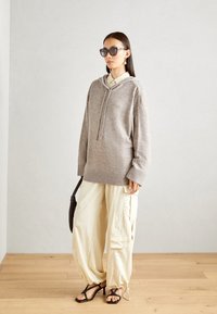 Sudadera de punto de gran tamaño en gris claro con cordón en el frente, combinada con pantalones cargo crema y sandalias negras con tiras. Accesorios incluyen gafas de sol.