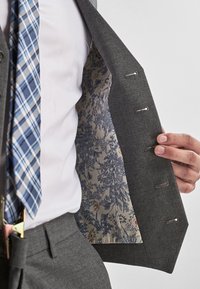 Graues Sakko mit einem floral gemusterten beigen Innenfutter. Das Sakko hat einen Reverskragen und vier Knöpfe auf der Vorderseite. Eine blau-karierte Krawatte wird getragen.