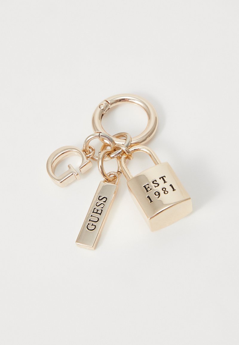 Porte-clés doré avec un cadenas portant la mention "EST 1981", une étiquette rectangulaire gravée "GUESS" et un fermoir circulaire. Texture métallique lisse.