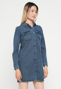 Tumši zila džinsas krekla kleita ar pogu aizdari, apkakli, garām piedurknēm un divām krūšu kabatām ar atloka aizdarēm.