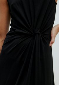 Schwarzes Kleid aus weich fließendem Stoff, mit einer gedrehten Knotendetail im Taillenbereich und drapiertem Stoff an der Seite. Glatte Textur und fließende Silhouette.