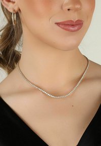 Silverfärgad halskedja med en böjd design, prydd med klara stenar som är arrangerade i en kontinuerlig rad. Slät yta, bärs mot en mörk topp.