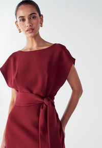 Viininpunainen mekko, joka on löysästi istuva ja jossa on leveä pääntie, varustettuna vyötärölle solmittavalla yksityiskohdalla. Sileä kangas, jossa on hieno ja elegantti rakenne.