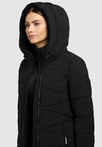 Schwarze gepolsterte Jacke mit Kapuze, Reißverschluss und diagonalen Nähten. Weiche Textur mit Seiten Taschen und markanten Akzenten.