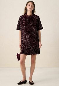 Robe bordeaux à sequins avec manches courtes, silhouette droite et col rond. Associée à des ballerines noires ornées et un petit sac.