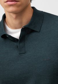 Dunkelgrünes Poloshirt aus strukturiertem Stoff, mit einem kragen, der geknöpft ist, und einer kontrastierenden weißen Innenschicht, die am Ausschnitt sichtbar ist. Minimalistisches Logodesign.