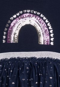 Laulu T-paita merivoimansinisellä värillä, jossa on paljeteista tehty sateenkaaren malli violetilla, hopeisella ja kultaisella. Paidassa on kimaltava vyötärönauha ja frilla-tüllihame sydänkuvioilla.