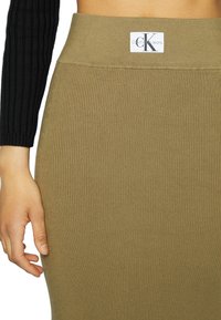 Olivgrön ribbad kjol med en bred midjeband som har en Calvin Klein Jeans-logotag. Materialet är elastiskt med en texturerad yta.