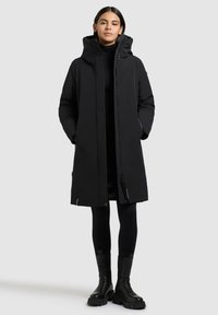 Noor, kes kannab pikka musta kapuutsiga talvekasukat, musti retuuse ja volüüme musti saapaid, seisab käed mantli taskutes.