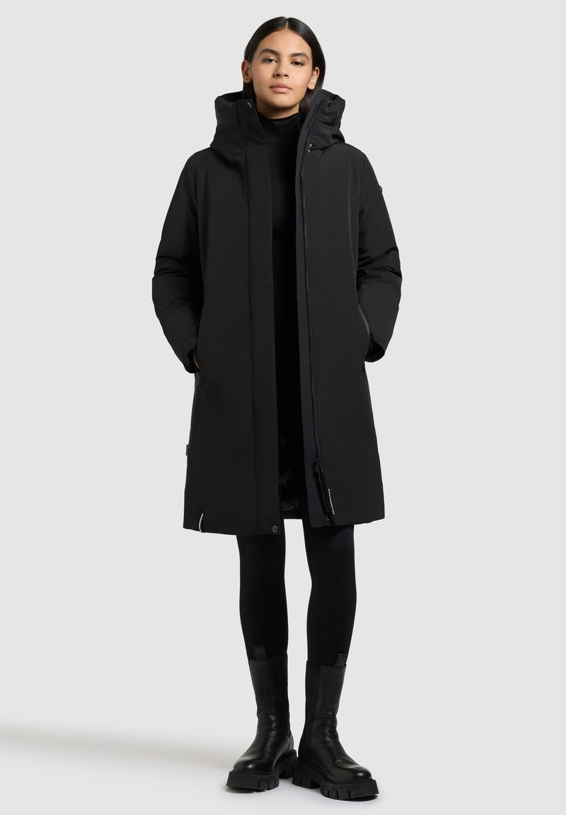 Noor, kes kannab pikka musta kapuutsiga talvekasukat, musti retuuse ja volüüme musti saapaid, seisab käed mantli taskutes.