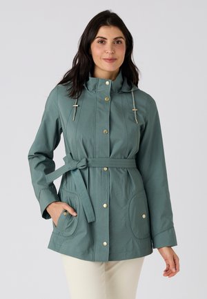 Femme portant une veste ceinturée bleu sarcelle avec des boutons dorés, main dans la poche, debout devant un fond uni gris clair.
