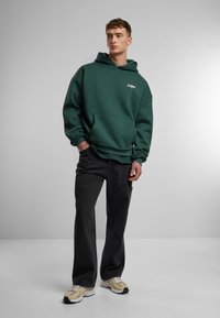 Sudadera verde con un bolsillo frontal y logo blanco, combinada con jeans negros y zapatillas beige, de pie frente a un fondo gris claro.