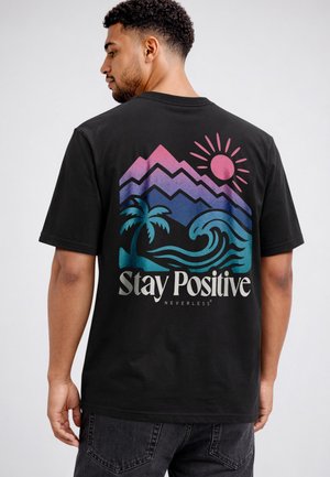 Mann trägt schwarzes T-Shirt mit farbigem Berg-, Sonne-, Palmen- und Wellenmotiv und dem Schriftzug „Stay Positive“ auf dem Rücken.
