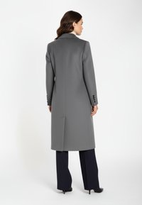 Langer grauer Mantel mit strukturiertem Muster, einfachem Schlitz und schwarzen Knöpfen, kombiniert mit marineblauen Hosen und schwarzen High Heels. Rückansicht gezeigt.