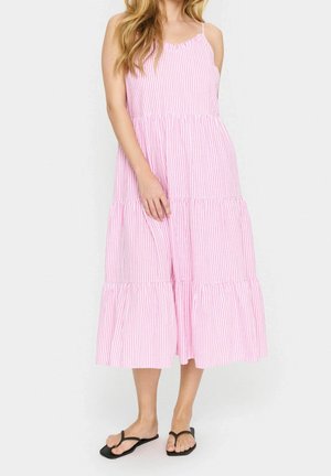 Frau trägt ein ärmelloses Midi-Kleid mit pink-weißen Streifen und einem gestuften Rock sowie schwarze flache Sandalen, steht vor einem schlichten Hintergrund.