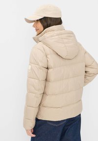 Beige dunjakke med hætte, der har quiltede sektioner og en glat overflade. Båret sammen med en lys beige kasket og mørkeblå jeans.