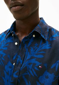 Camicia a maniche corte con bottoni di colore blu scuro, decorata con un motivo floreale di un blu più chiaro. Presenta bottoni, colletto e un piccolo dettaglio con logo.
