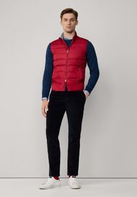 Chaleco acolchado rojo con cierre de cremallera, llevado sobre una camisa de manga larga azul marino. Modelo combinado con pantalones negros y zapatillas blancas.